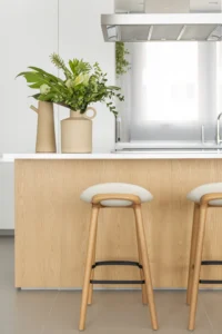 Oak kitchen island with a white top, upholstered stools and ceramic vases of foliage at Apartamento AM