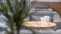 Two Basics Coffee cups on a round oak table in front of a grey quilted sofa, framed by foliage in the foreground