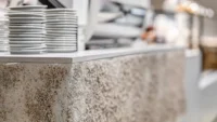 Close-up of the bush-hammered stone counter edge at Basics Coffee, with stacked saucers and the espresso machine behind