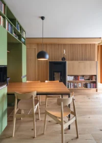 Dining area in the Cottage looking toward the fluted timber wall, with a black wood-burning stove and a low bookcase