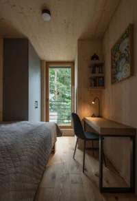Bedroom in the Cottage lined in pale spruce boards, with a compact desk, a reading lamp and a full-height window to the trees