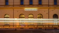Illuminated MASARYČKA sign on the historic station facade at night, with tram light trails streaking past the arched windows