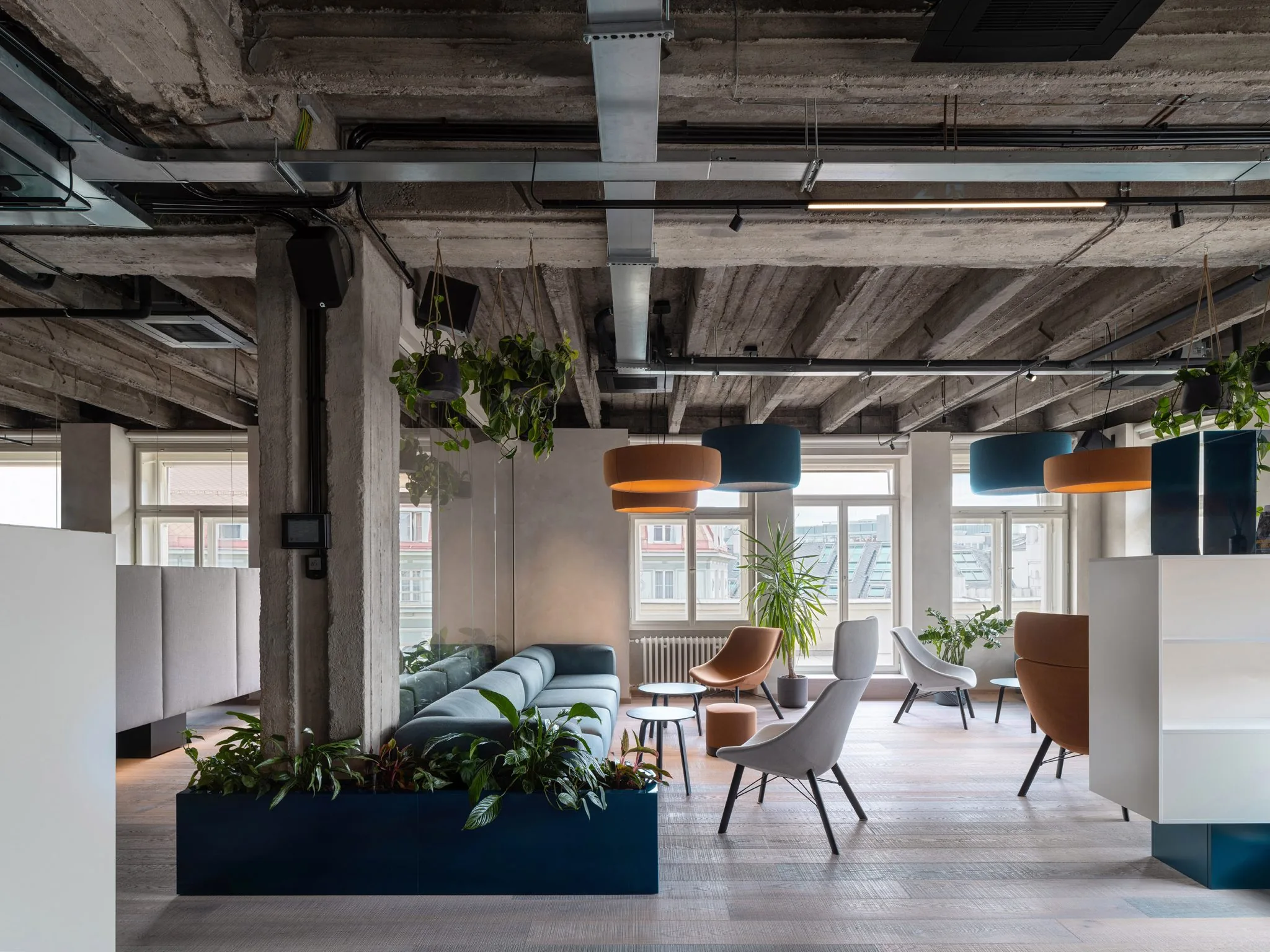 Open work area at Office That Breathes, with an exposed concrete ribbed ceiling and services, a blue planter box behind a long sofa, and grey acoustic screens dividing the floor