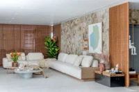 Long white sofa set against a stone wall between timber muxarabi screens, Apartment Curitiba by Fernando Luiz Dal Bosco