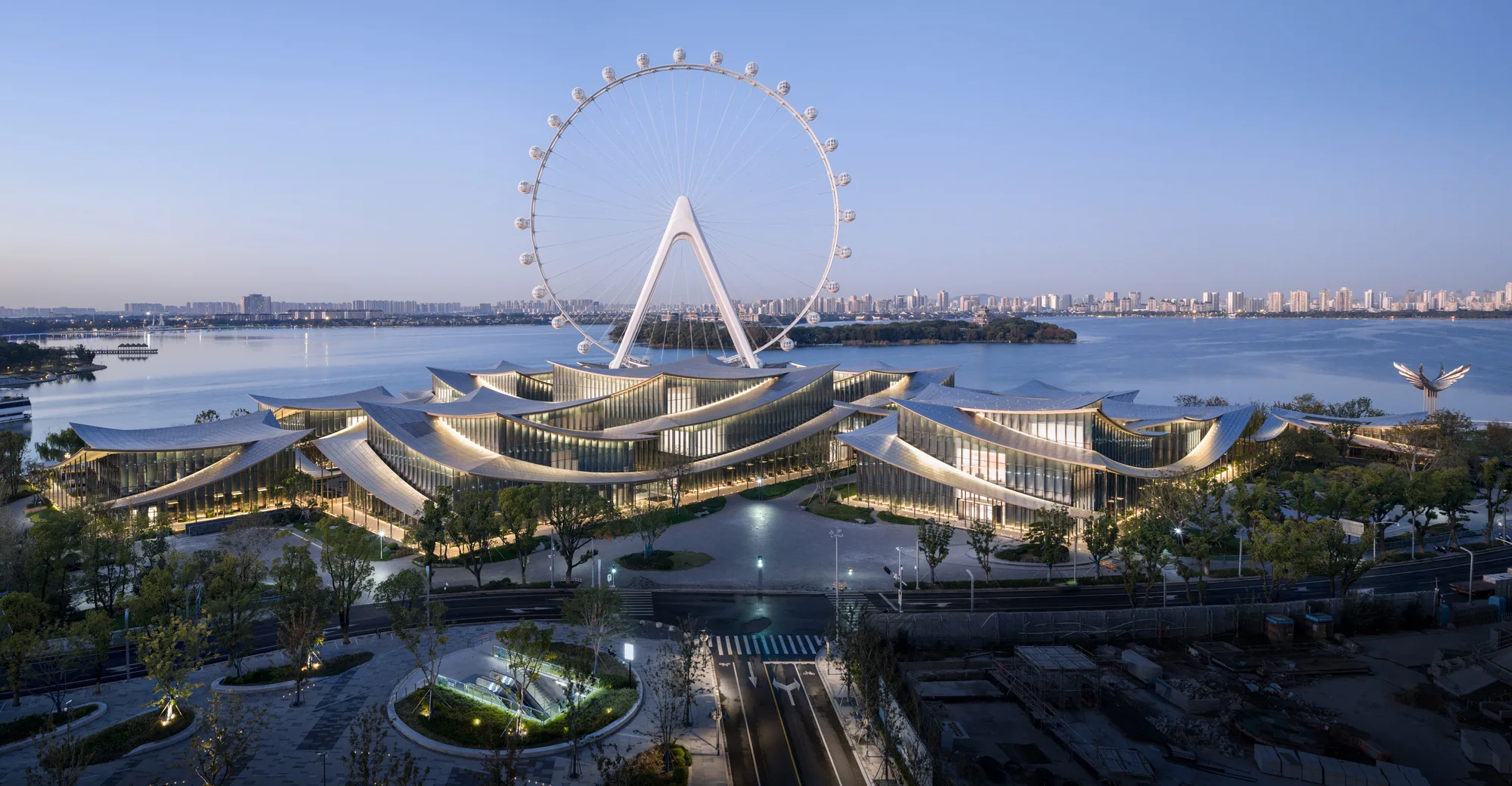 Wave-shaped low-rise museum complex illuminated at dusk beside a lake, with a large Ferris wheel behind it