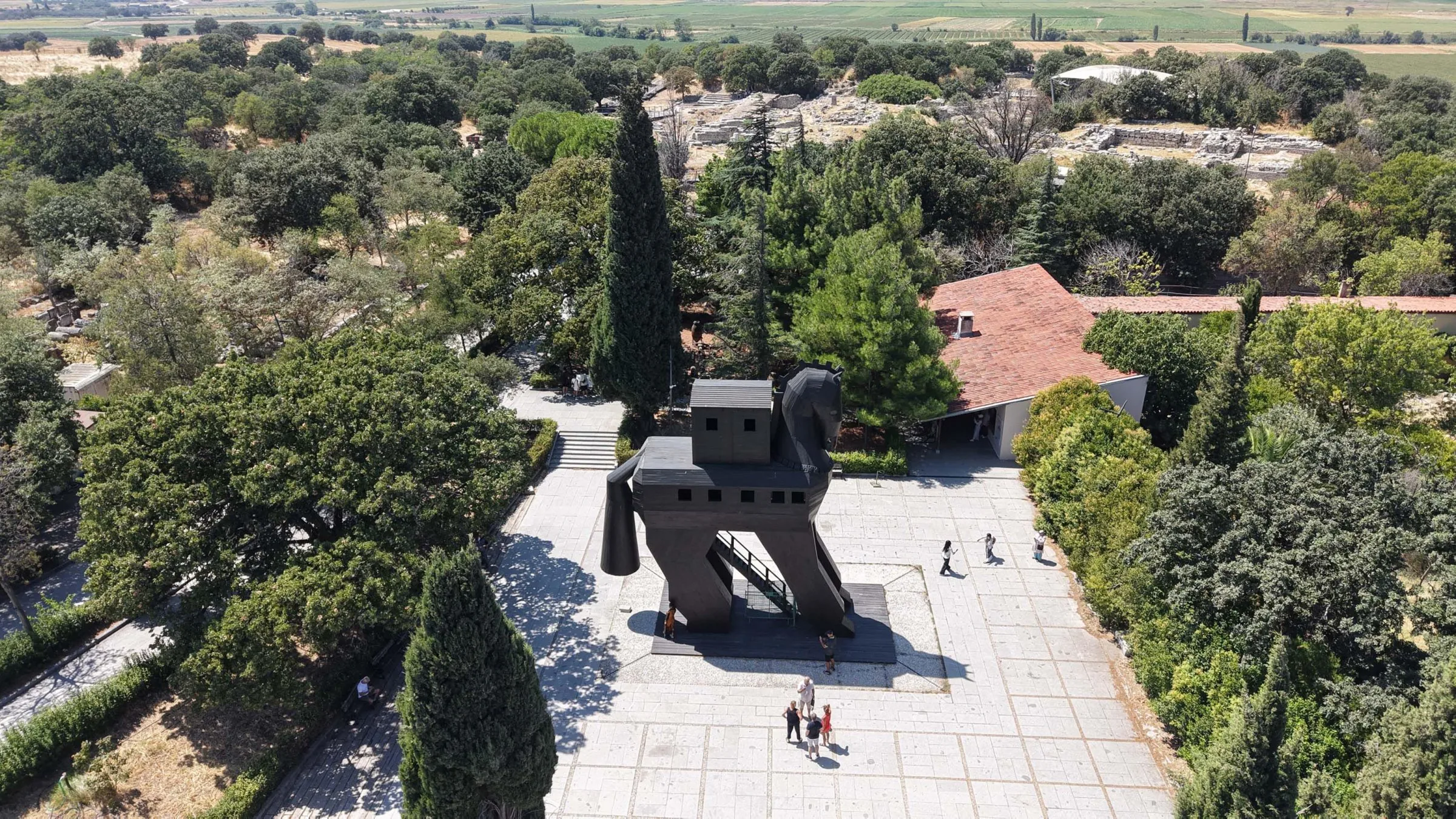 Aerial view of a large wooden horse statue on a paved plaza surrounded by trees near a tiled-roof building