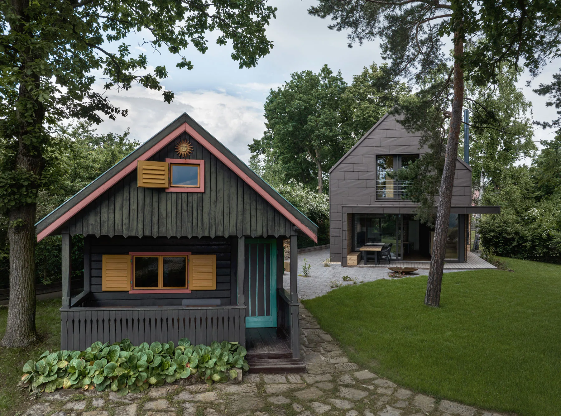 The original painted timber cottage with its carved porch and yellow shutters in the foreground, with the new dark-clad Cottage standing behind it