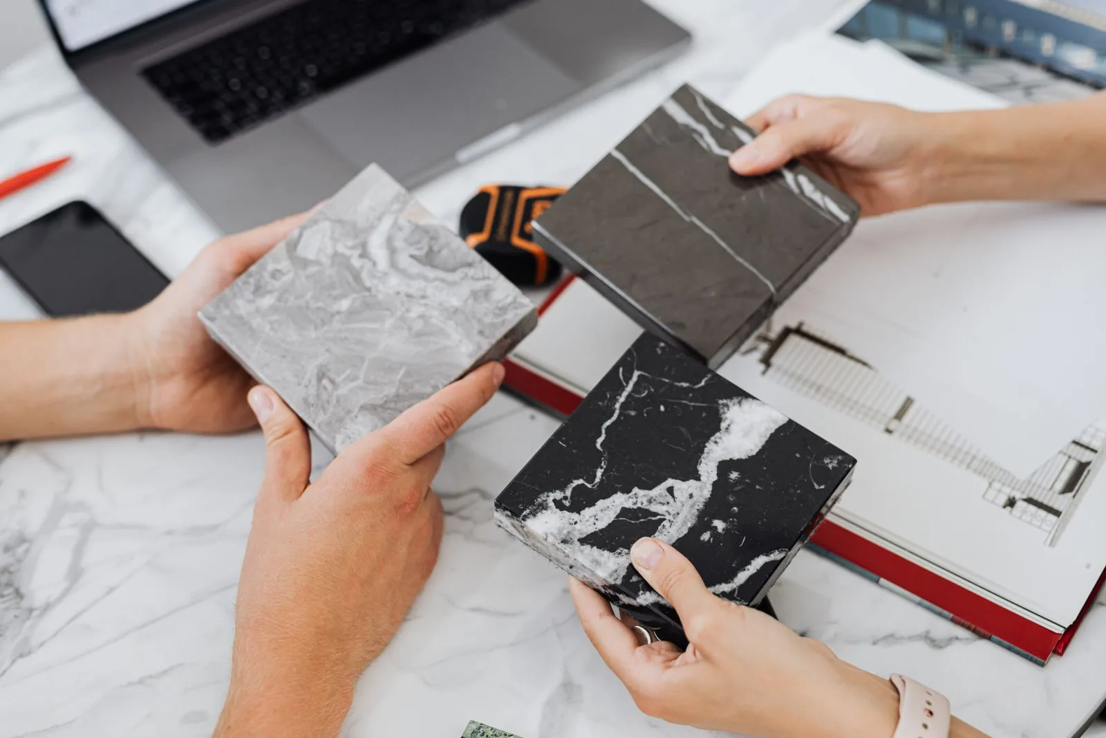 Hands holding grey and black marble samples over a printed floor plan and a laptop