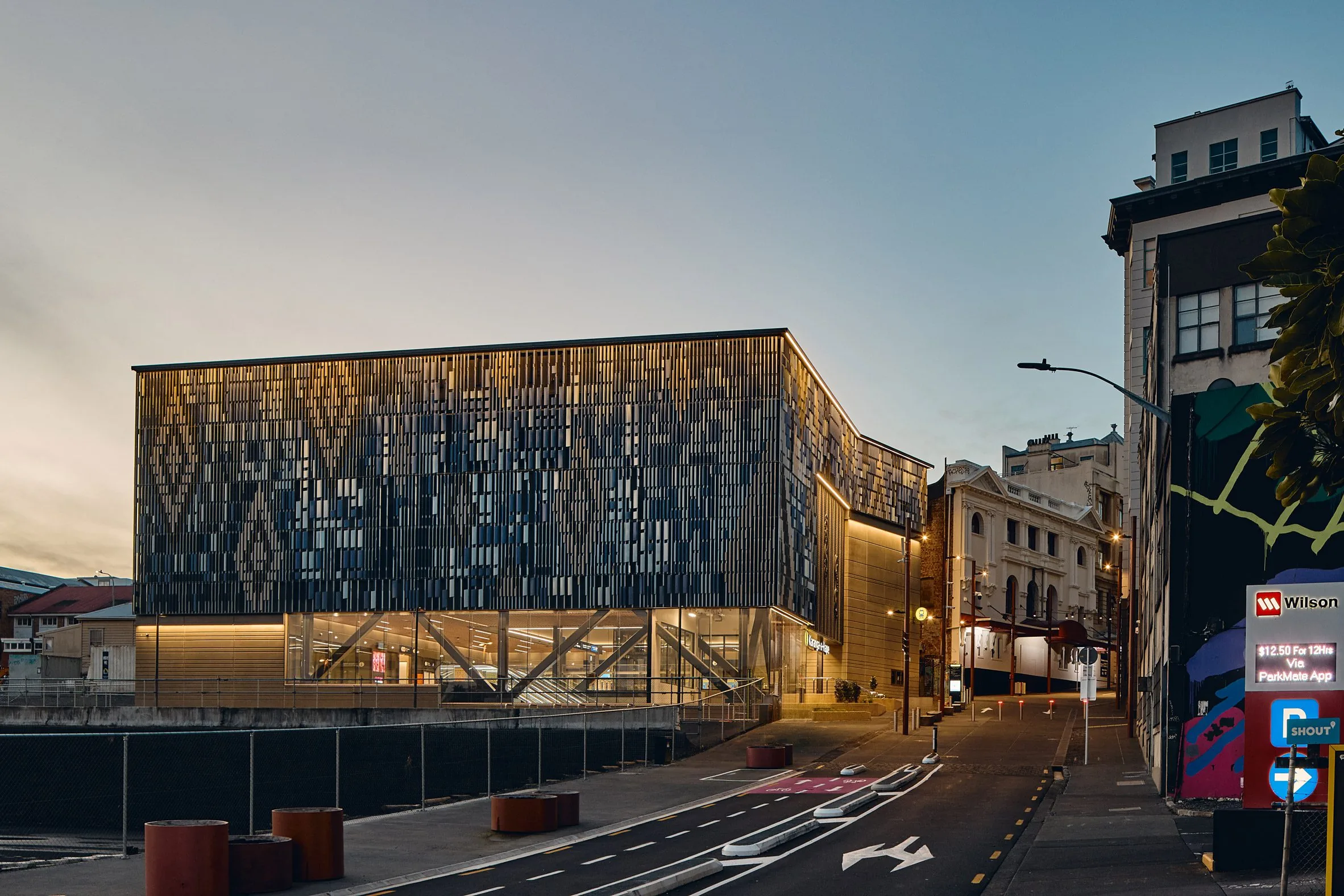 Rectangular building with a diamond-patterned metal facade illuminated at twilight beside older brick buildings
