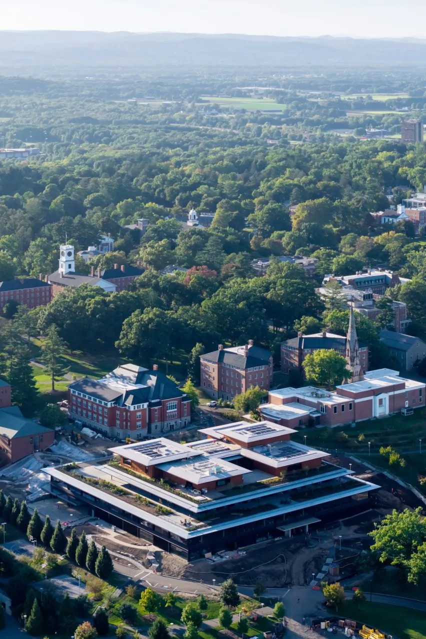 Wide aerial shot of a wooded campus with a modern flat-roofed building near historic brick halls and hills beyond