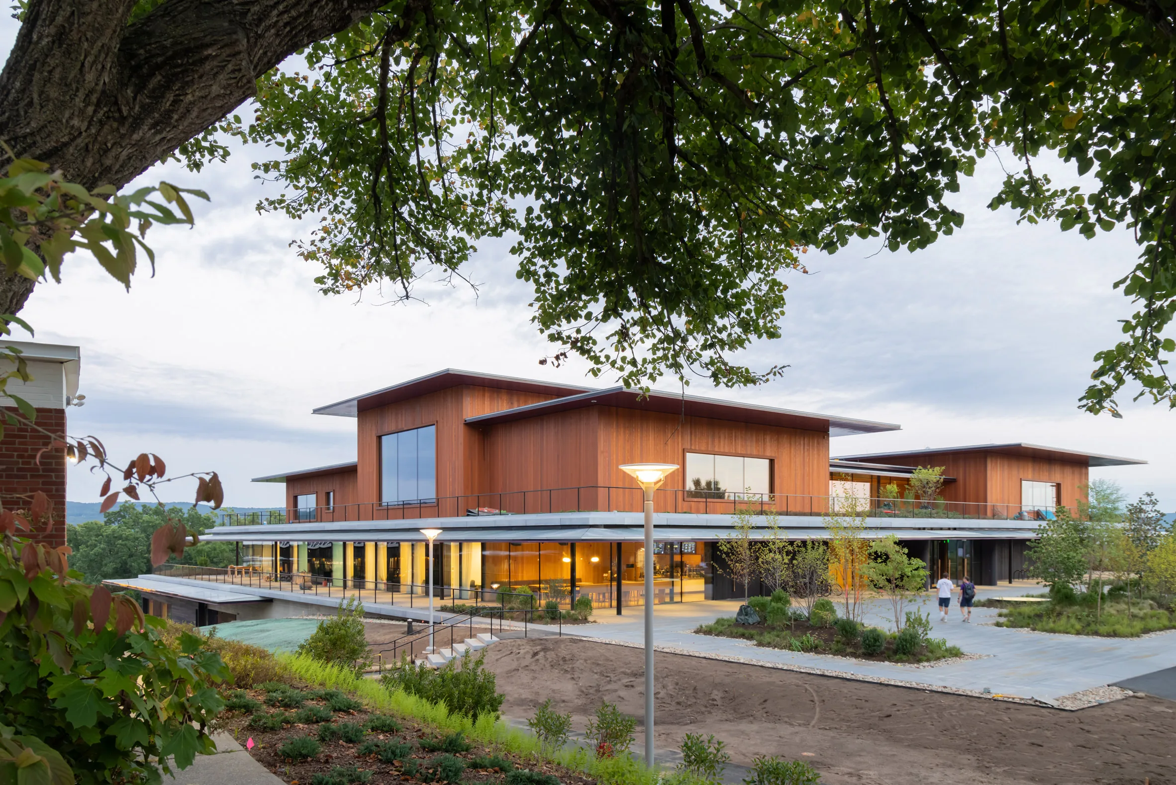 Wood-clad building with overhanging roof and glowing interior lights, seen through tree branches at dusk