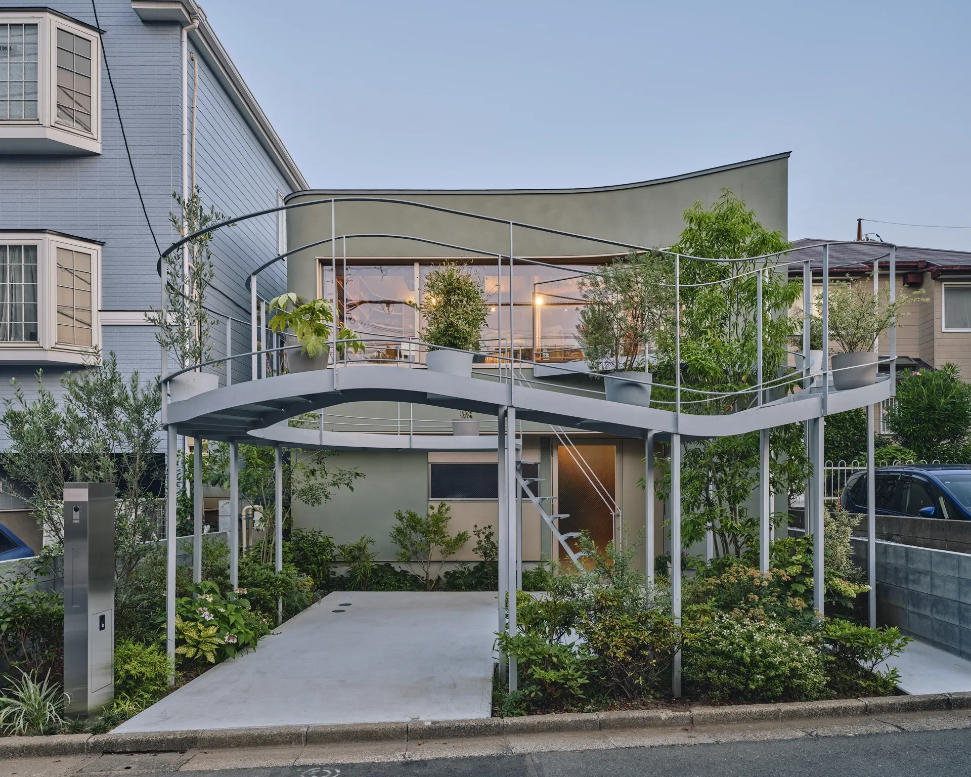 House in Nakano seen from the street at dusk with the curved planted terrace lit above the entrance