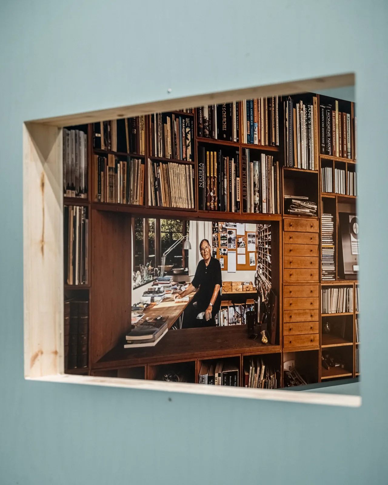 Rectangular opening in a pale blue wall framing a book-lined study with a person at a desk