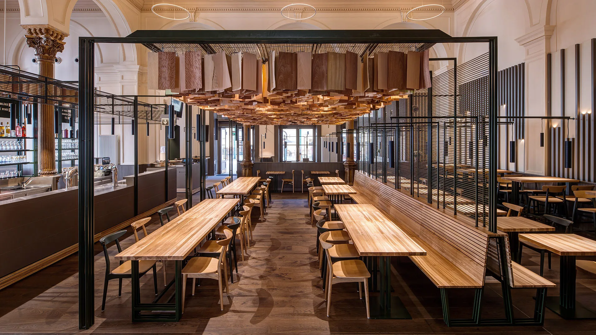 Central axis of Masaryčka Restaurant, with long communal tables and benches beneath the faceted timber ceiling cloud suspended in a black steel frame