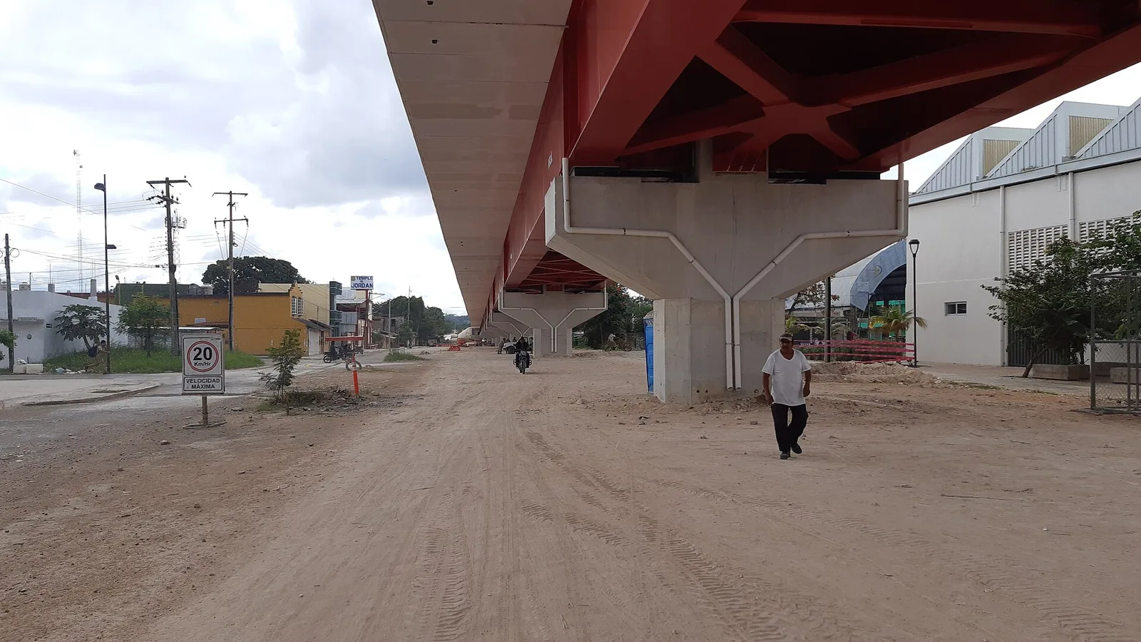 Underside of an elevated concrete railway viaduct above a dirt road with a pedestrian and low buildings