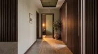 Corridor lined with dark timber slats leading to a doorway with a plant and olive plaster beyond