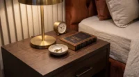 Dark timber nightstand with a brass lamp, clock and books beside a linen covered bed