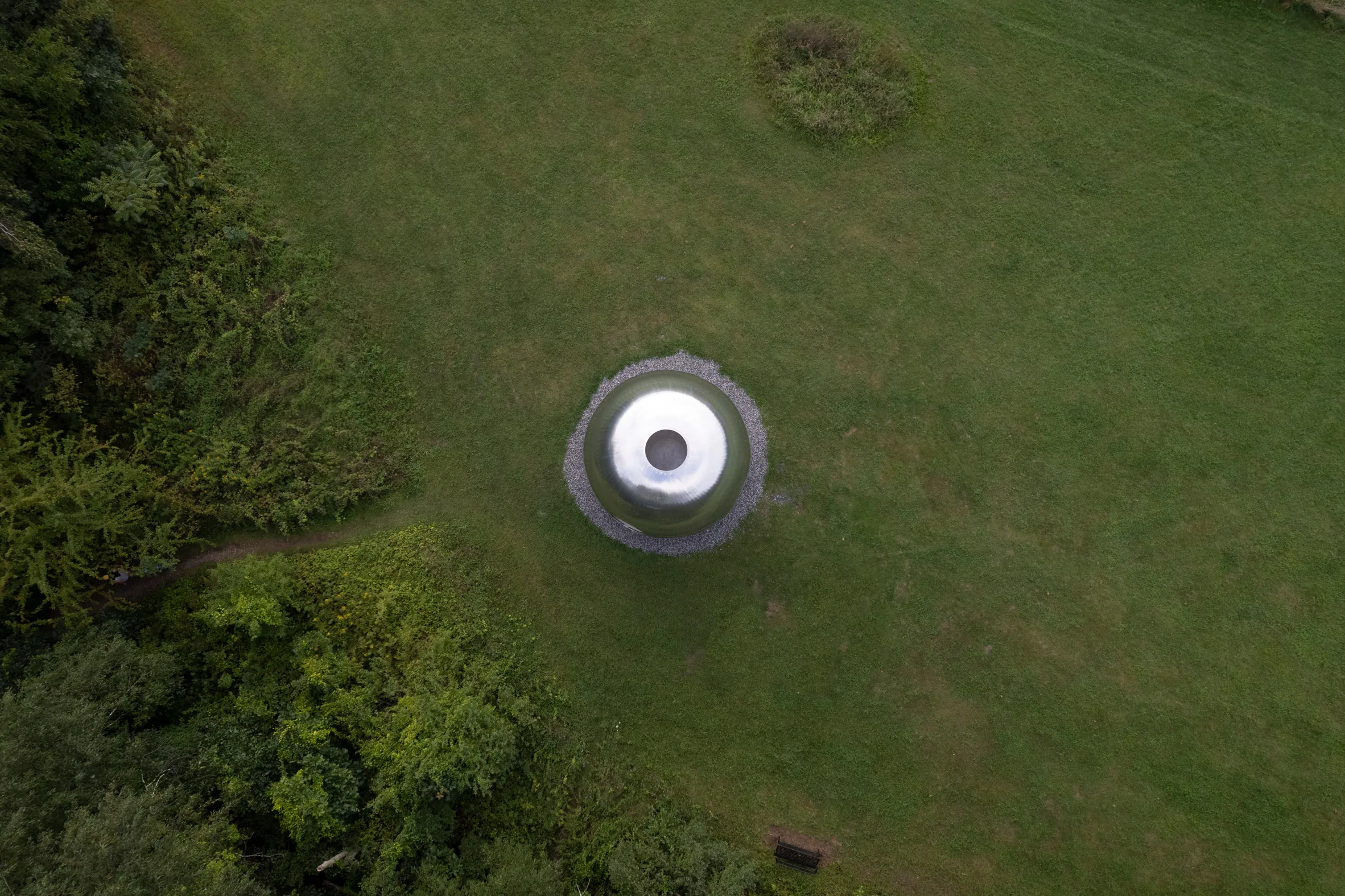 Aerial view of a round metallic structure with a circular opening set on a grassy lawn near trees