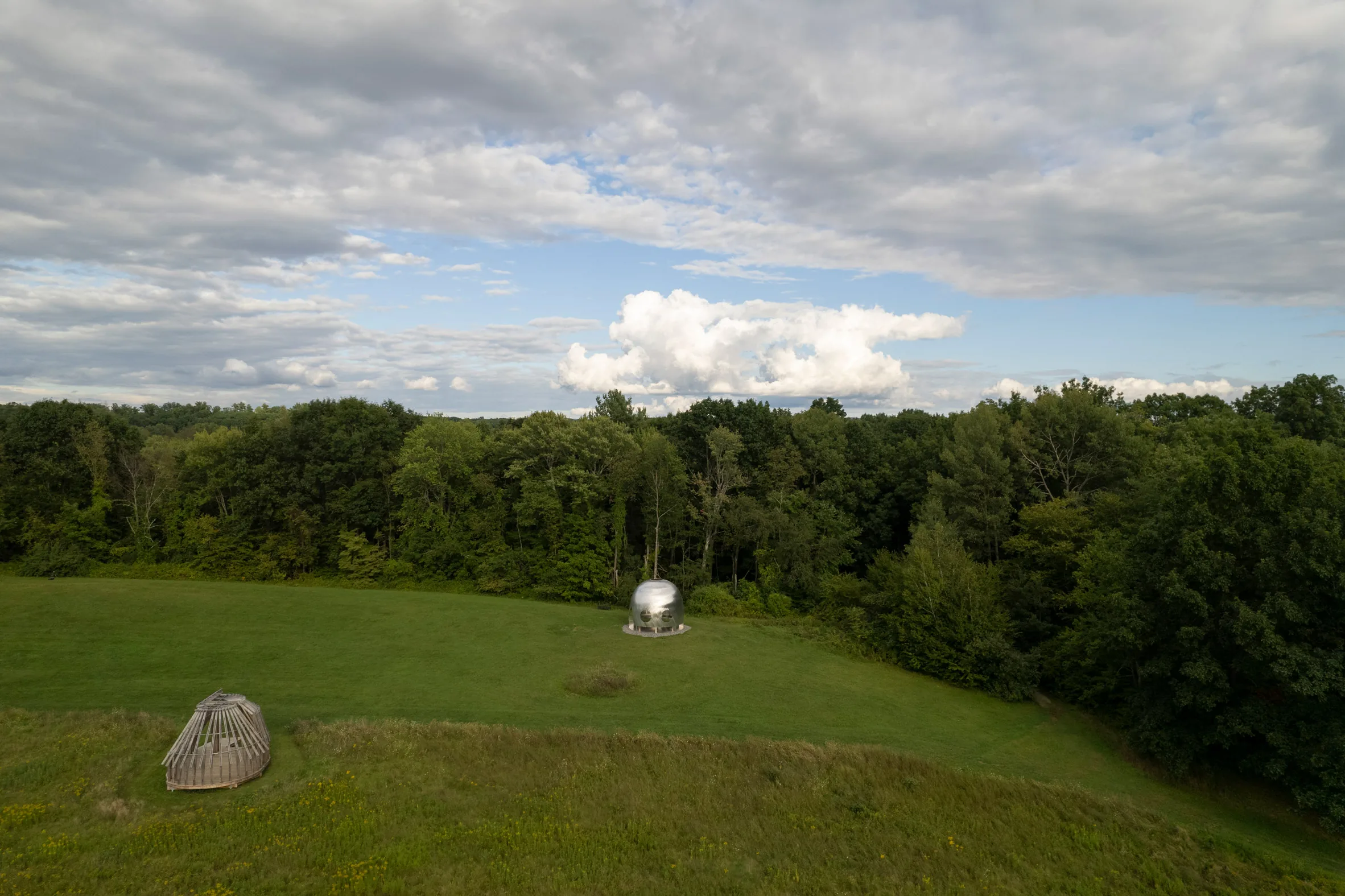 Wide aerial view of a grassy field with a small metallic dome and a wooden structure near forest edges under cloudy sky