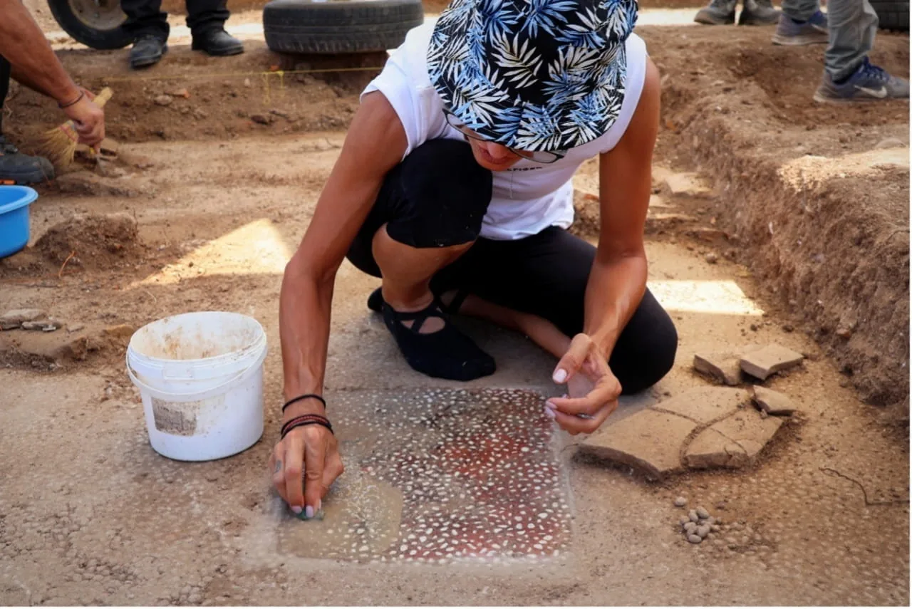 A person crouches on the ground, carefully brushing a patterned mosaic floor fragment beside a bucket