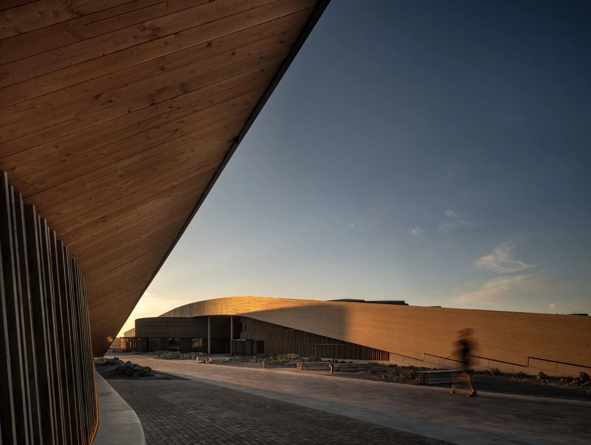 View from under a timber soffit toward the curved library volume across a brick paved forecourt