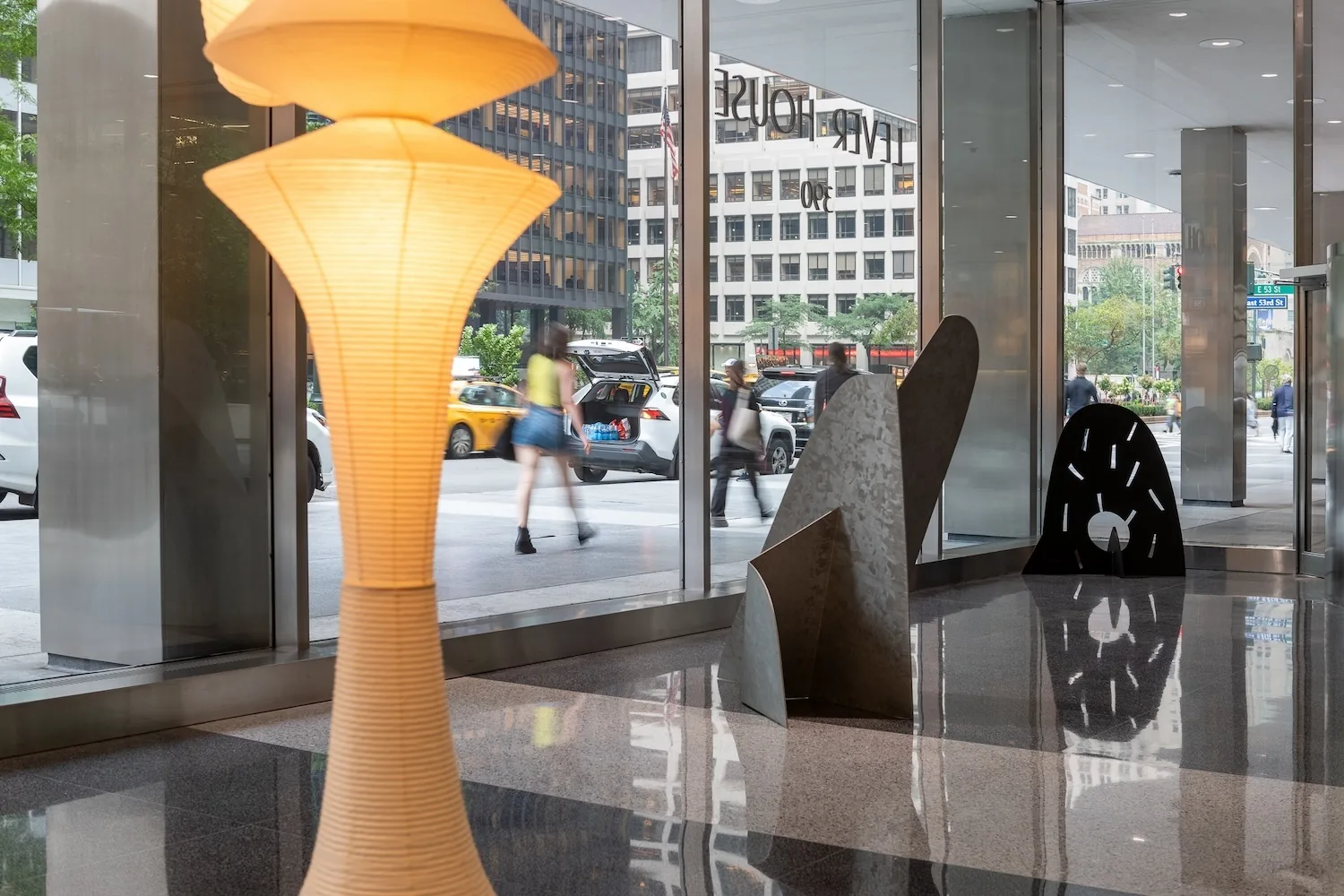 Glowing paper floor lamp beside angular metal sculptures in a glass-walled lobby overlooking a street