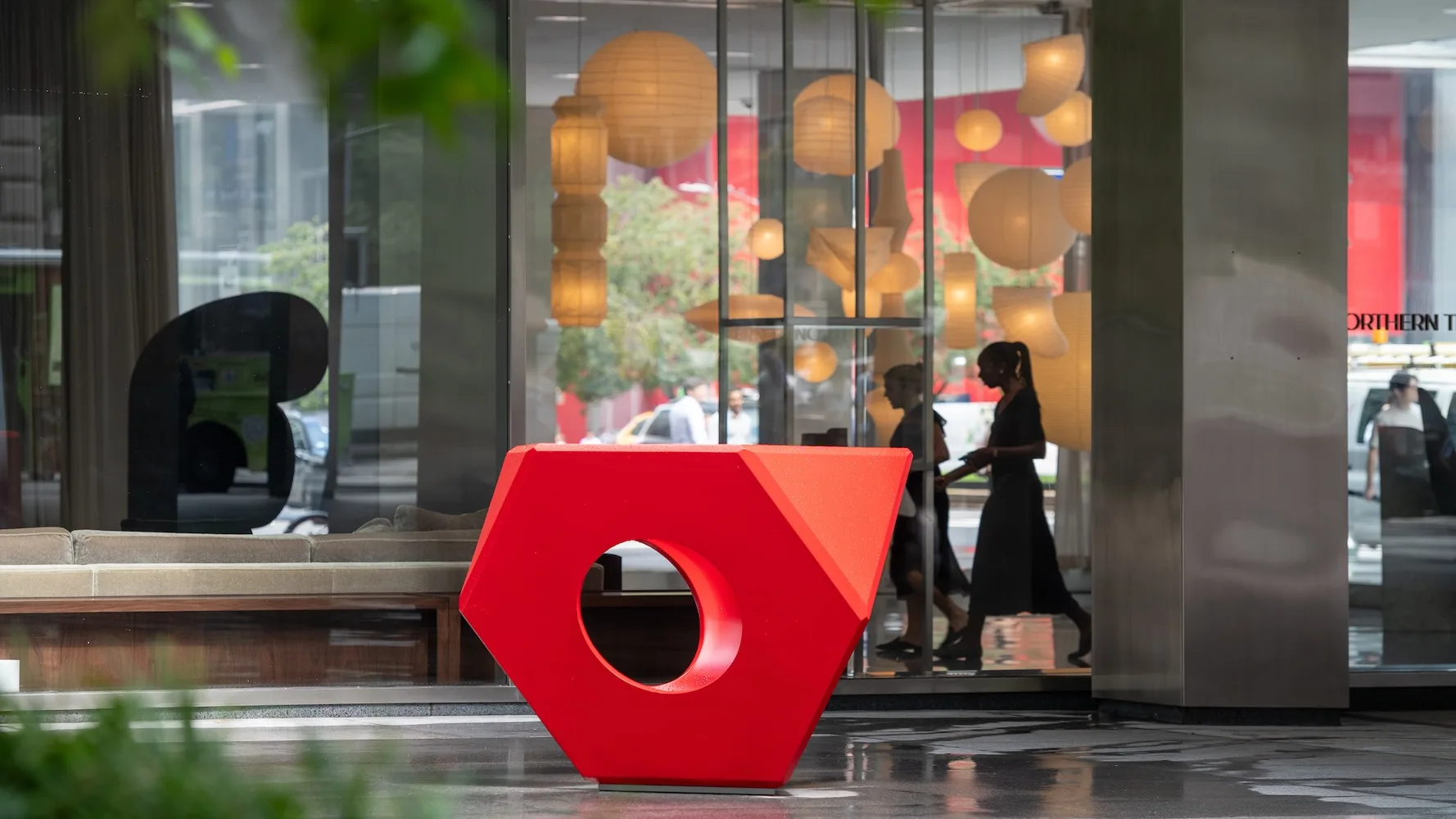 Large red hexagonal sculpture with a circular hole stands on a plaza near a glass building lobby