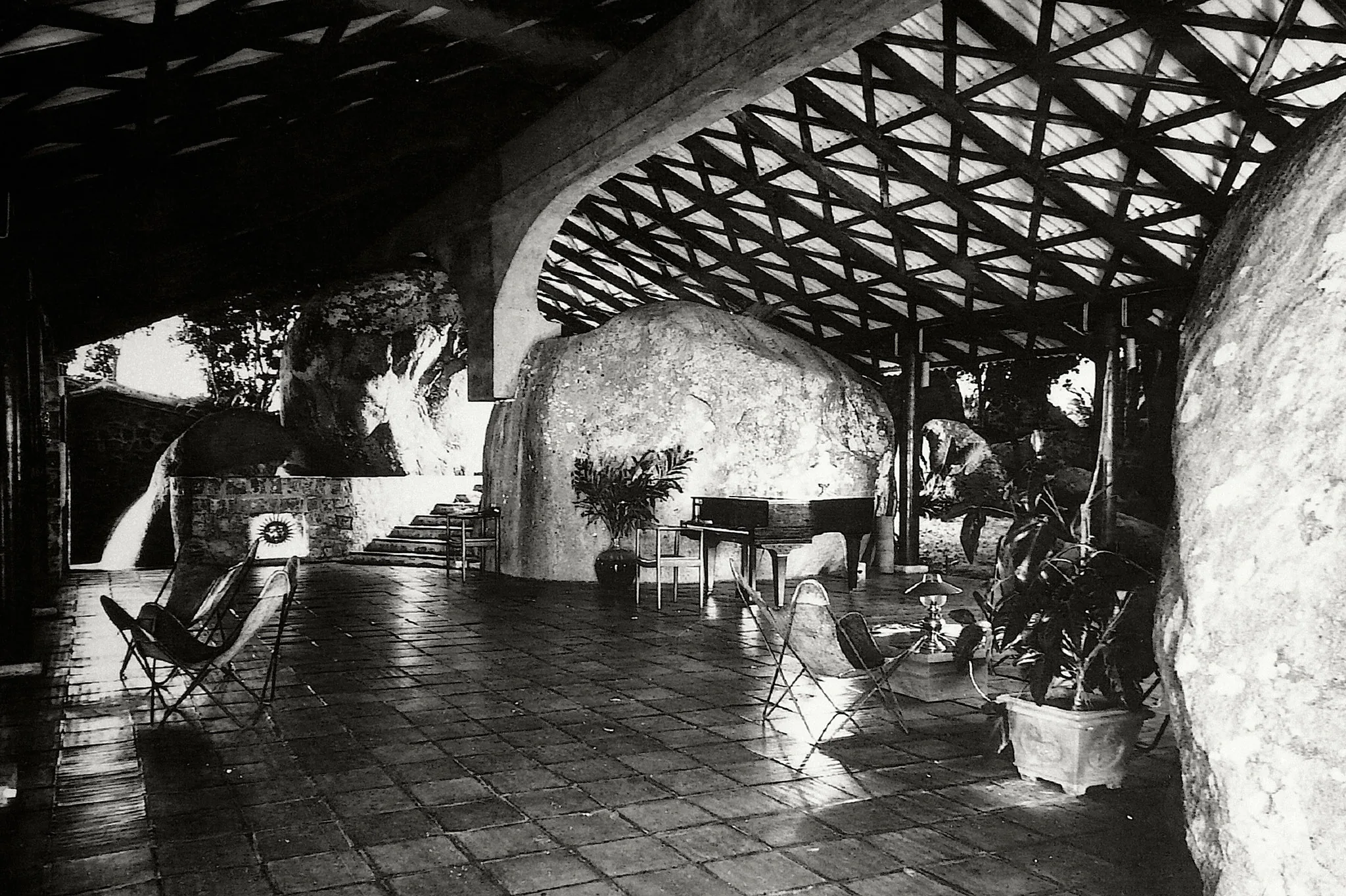 A covered stone terrace with a lattice roof, folding chairs, a piano and large natural boulders integrated into the