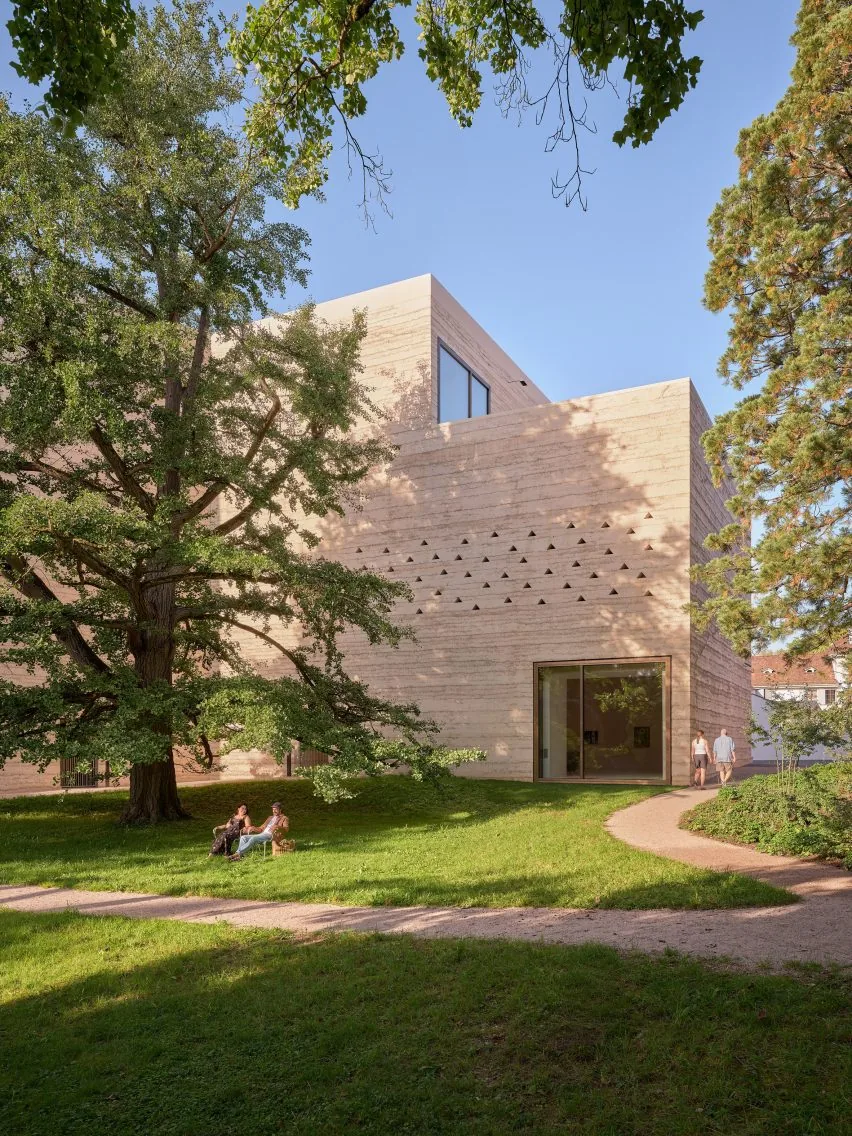 Corner of a textured stone building with small triangular openings, shaded by large trees