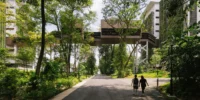 Two people walking along a tree lined road under an elevated timber clad bridge, Singapore Institute of Technology by Henning Larsen