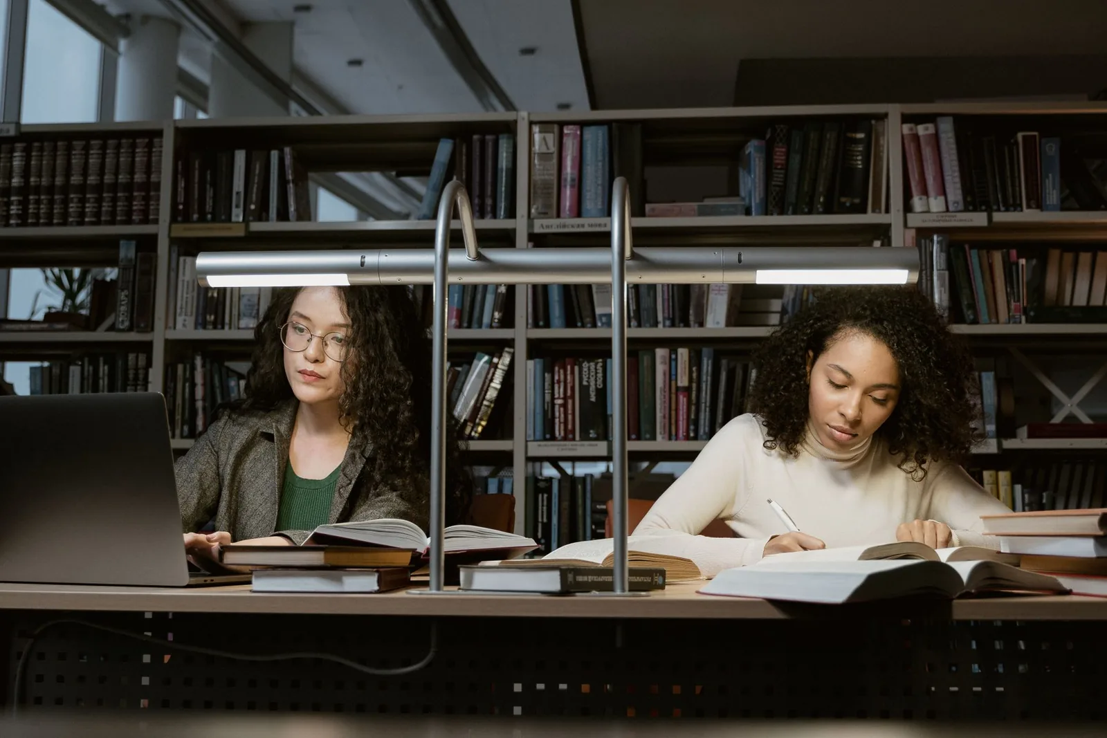 Two students reading and taking notes at a library desk surrounded by shelves of books