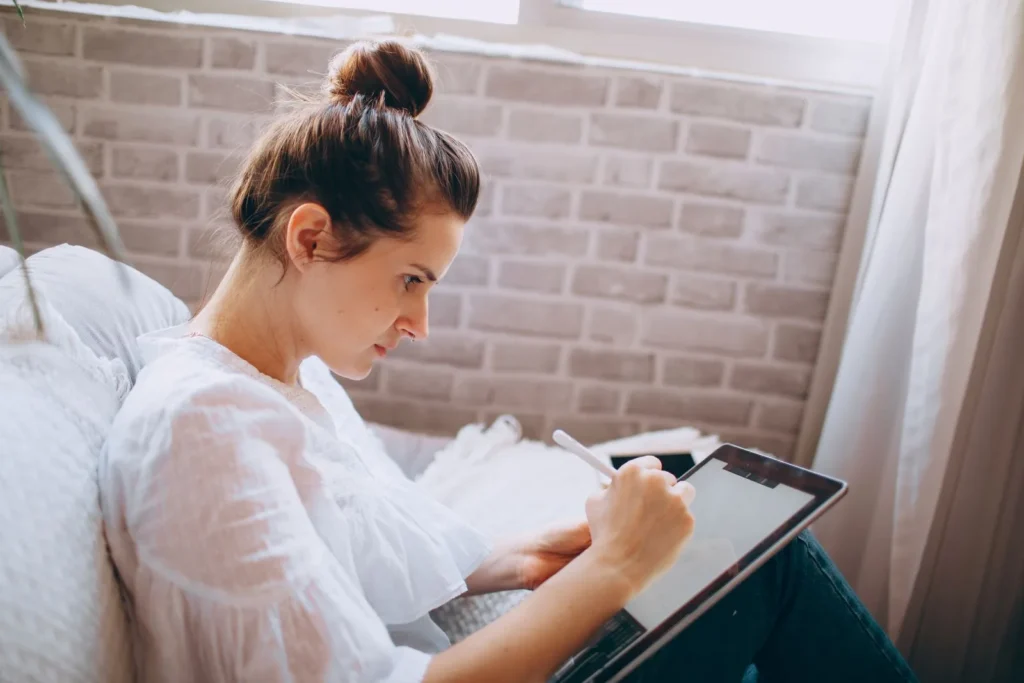 Woman sketching on a tablet with a stylus while sitting on a sofa at home, one of the ways mobile drawing apps fit into everyday practice
