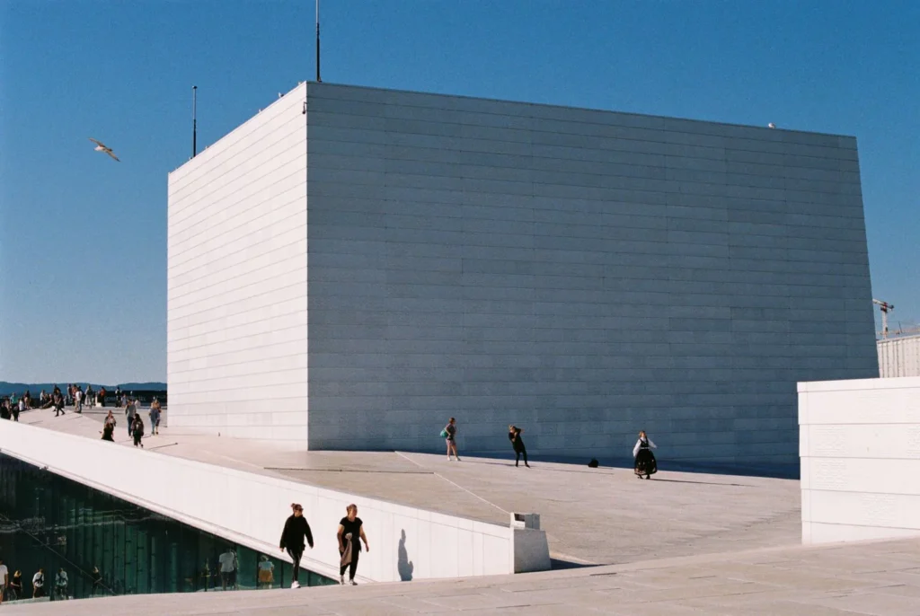 People crossing the marble roof of the Oslo Opera House beside its white stage tower, their small figures setting the scale