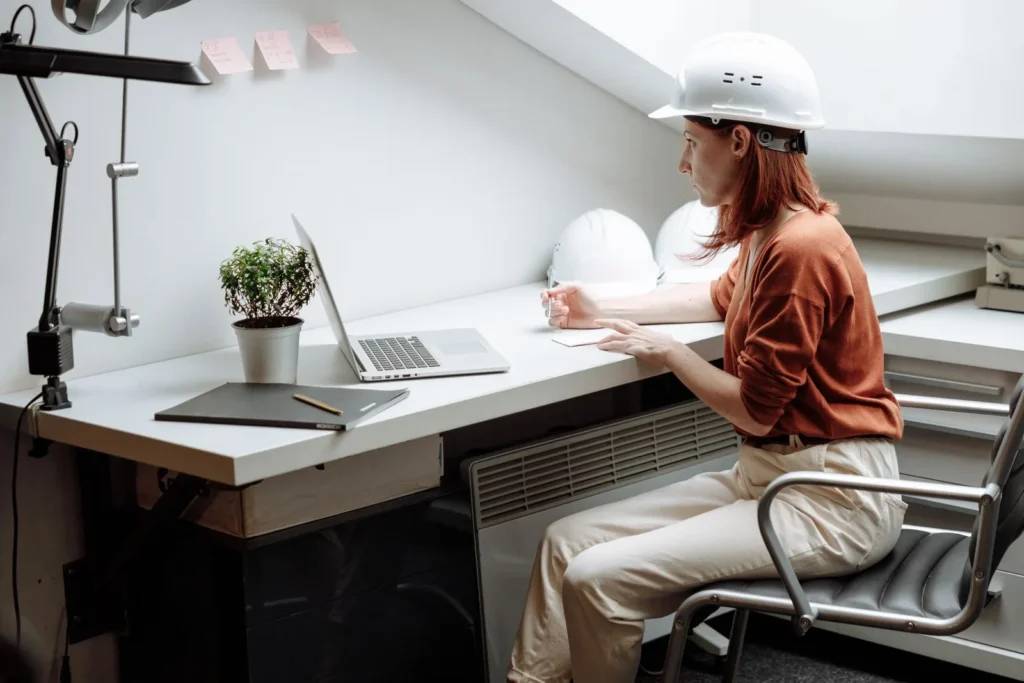 Architect in a white hard hat working on a laptop at a long studio desk, a typical setup for digital tools for architects