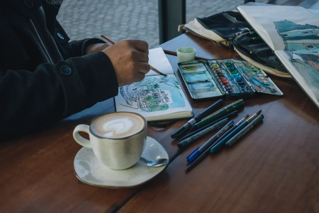 Sketching a street scene in a notebook with watercolors and pens beside a coffee in a cafe