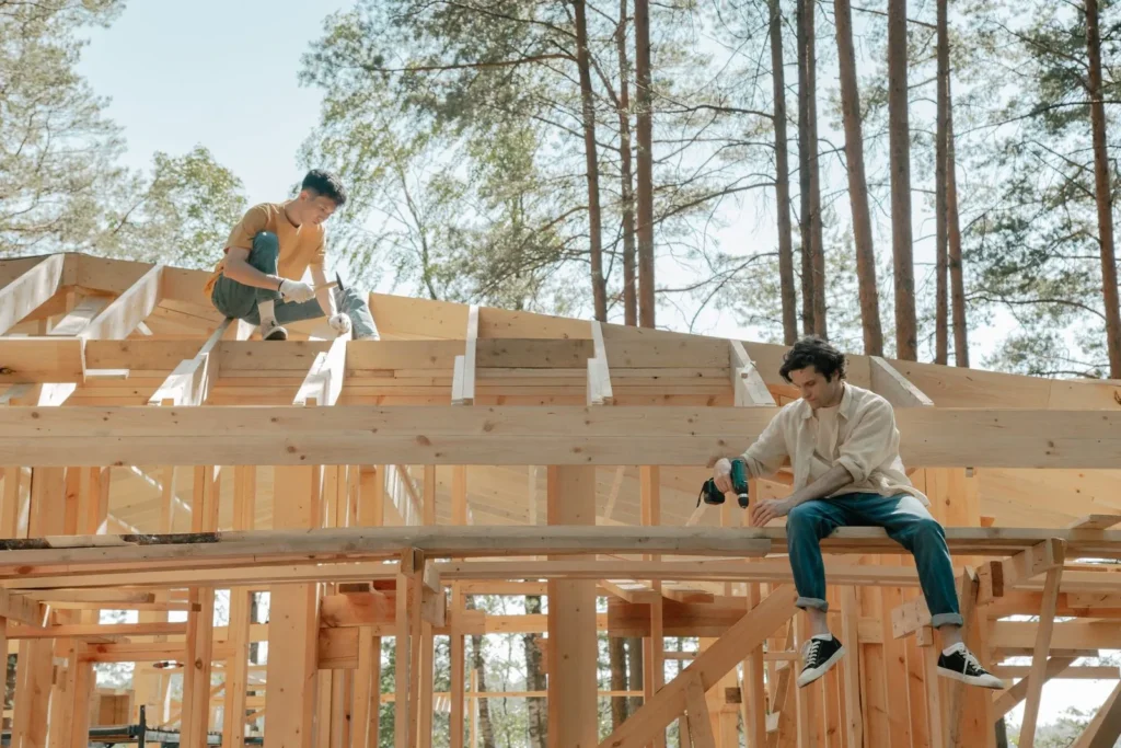 Two builders working on the timber frame of a house among pines, the kind of self-build saga TV series for architects follow