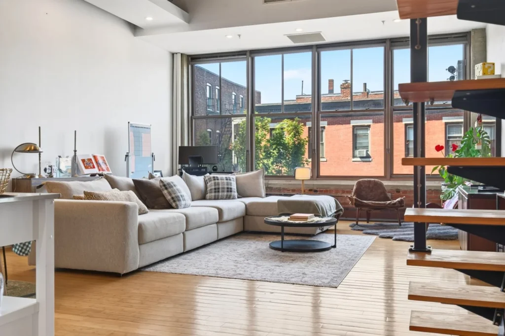 Open-plan loft with tall steel-framed windows, a sectional sofa and a floating stair in a former industrial building