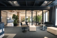 Communal lounge with low poufs and armchairs leading to a glazed garden kitchen with plants, ADDITIVE HQ by NOA