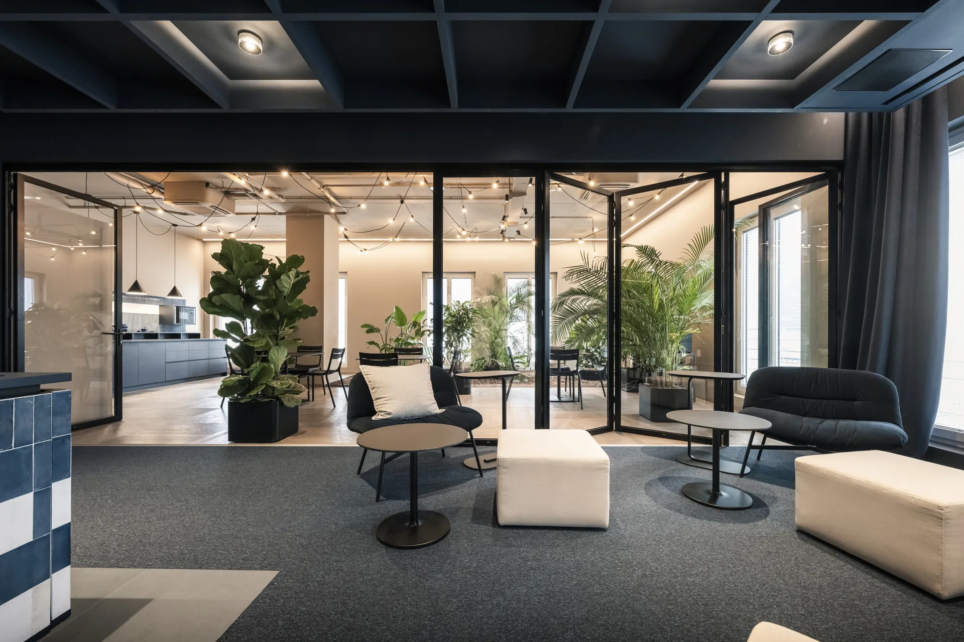 Communal lounge with low poufs and armchairs leading to a glazed garden kitchen with plants, ADDITIVE HQ by NOA