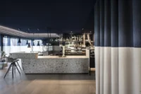 Bar with a terrazzo counter and backlit bottle shelves beside a curtain split into beige and blue, Aeon by noa*