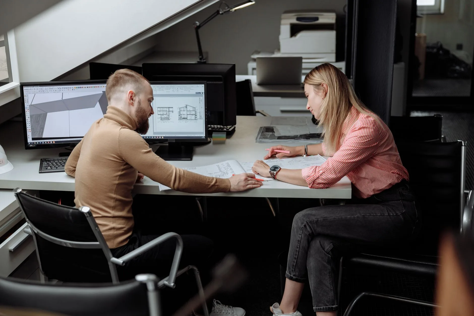 Two architects at a shared desk review printed drawings beside monitors showing a 3D model and building sections