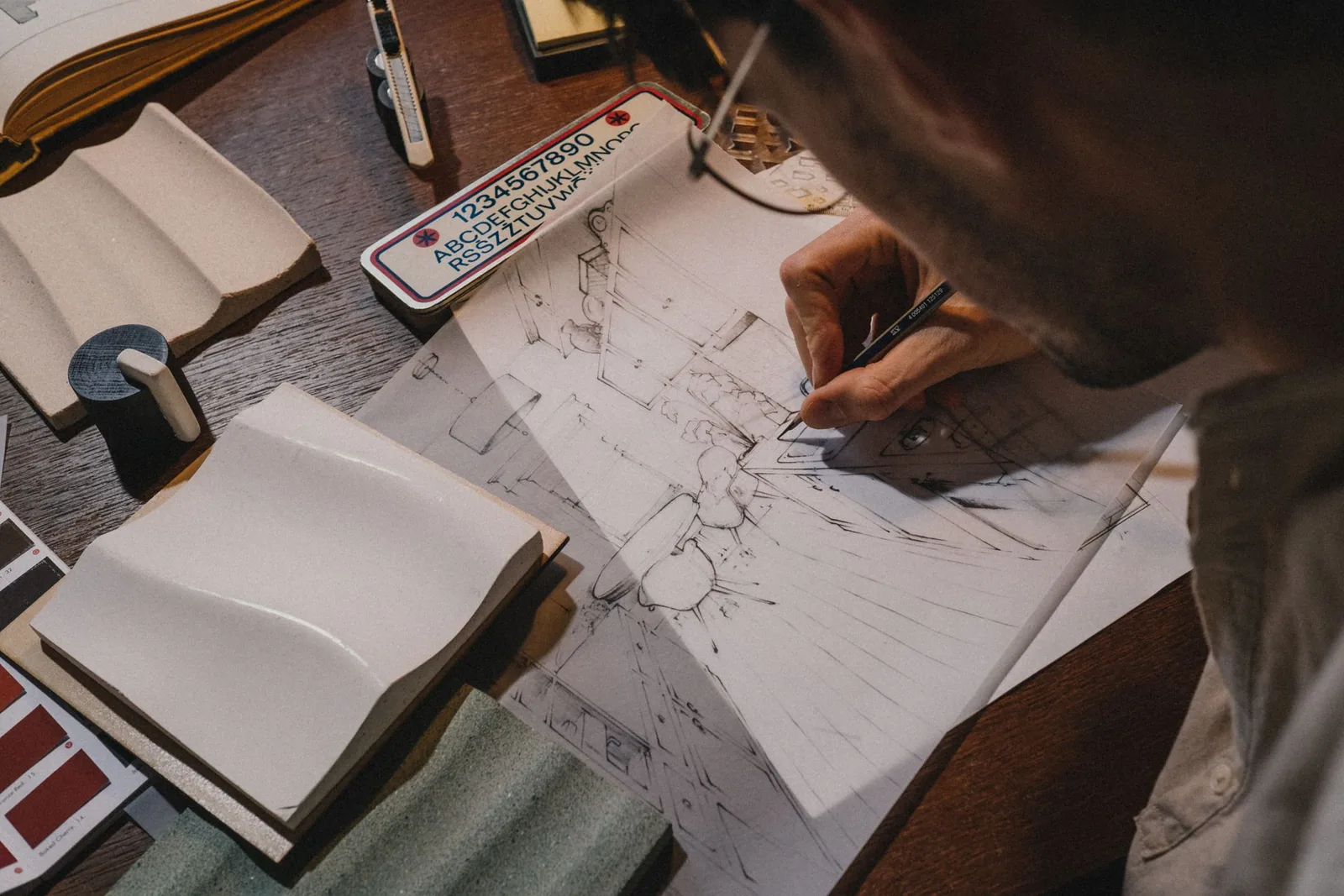 Architect sketching an interior perspective in pen on tracing paper beside material samples and a lettering stencil