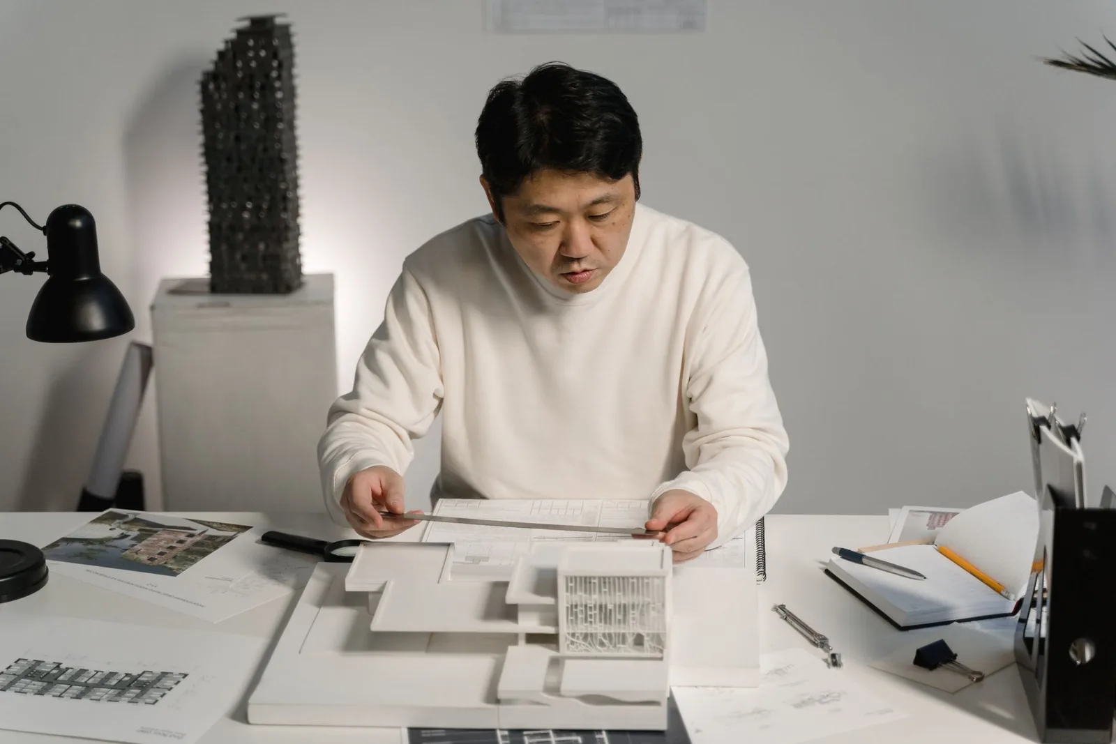 Architect measuring a white massing model on a desk covered with drawings and reference photos