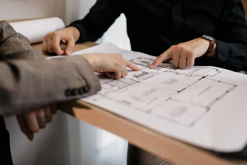 Two people pointing at details on a printed plan spread across a table