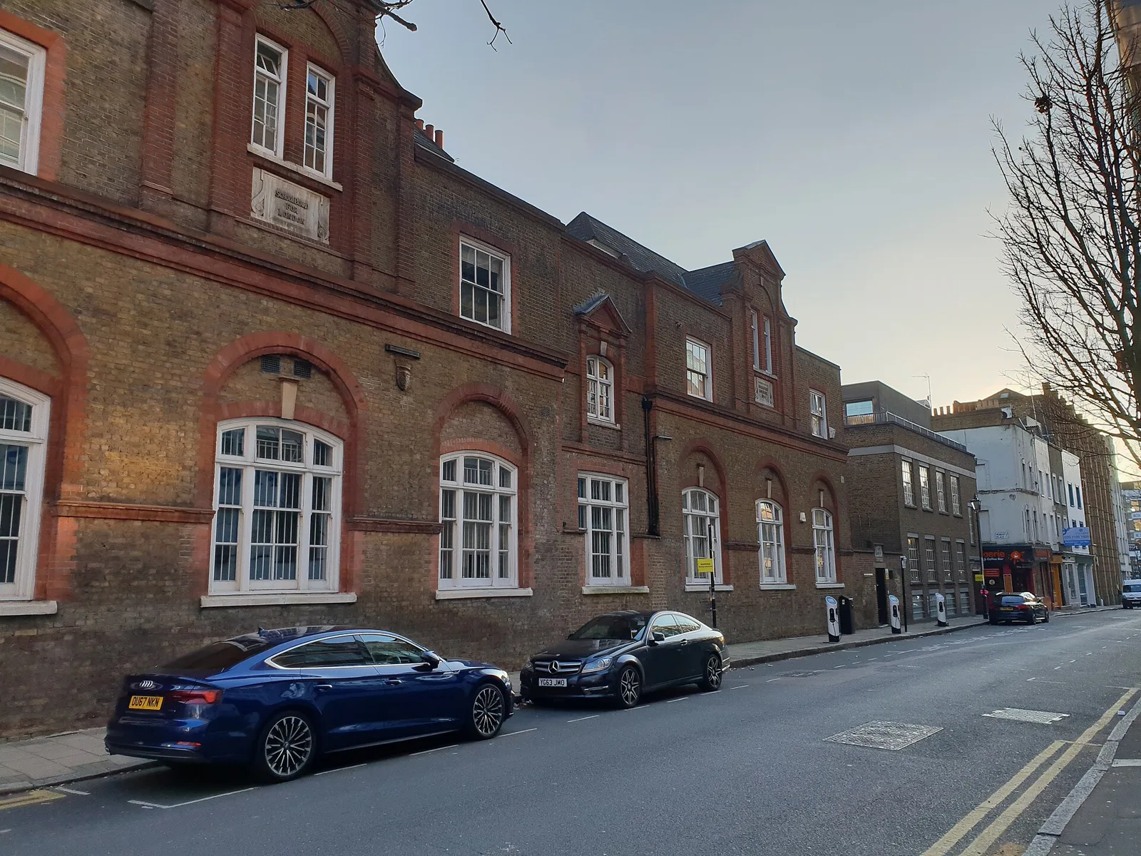 Long brick facade with rows of white sash windows and arched heads along a quiet street with two parked cars