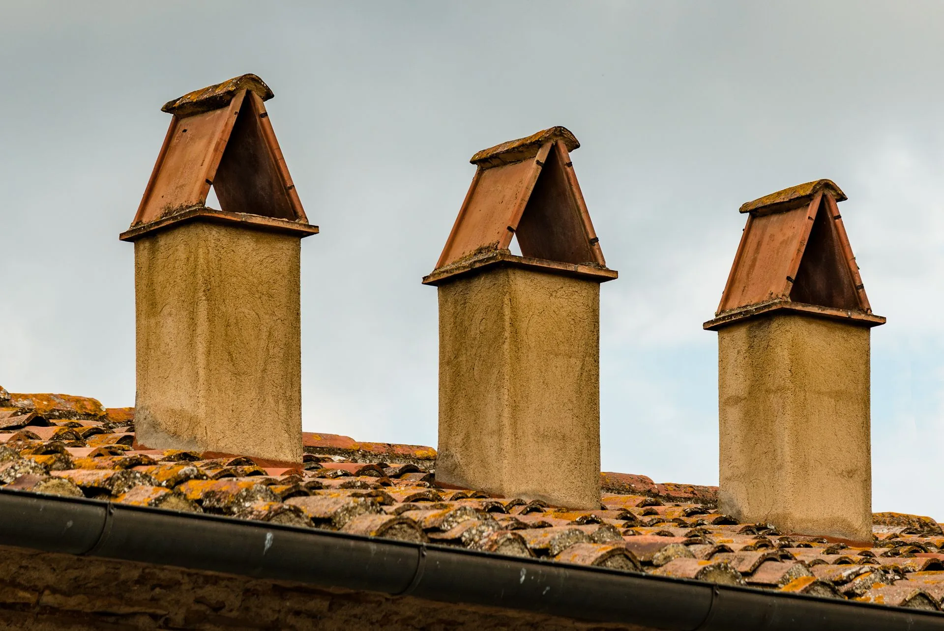 Three rendered chimney stacks fitted with triangular clay caps on a weathered tiled roof