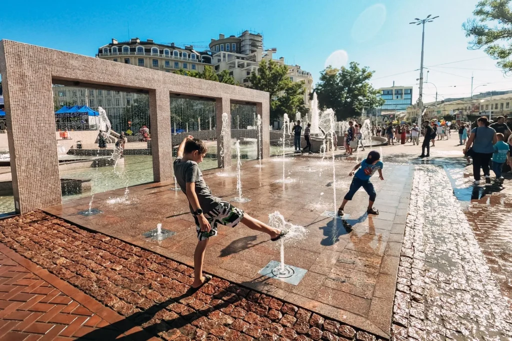 Children playing among ground level water jets in a sunny paved plaza, a lively example of experiential urban spaces