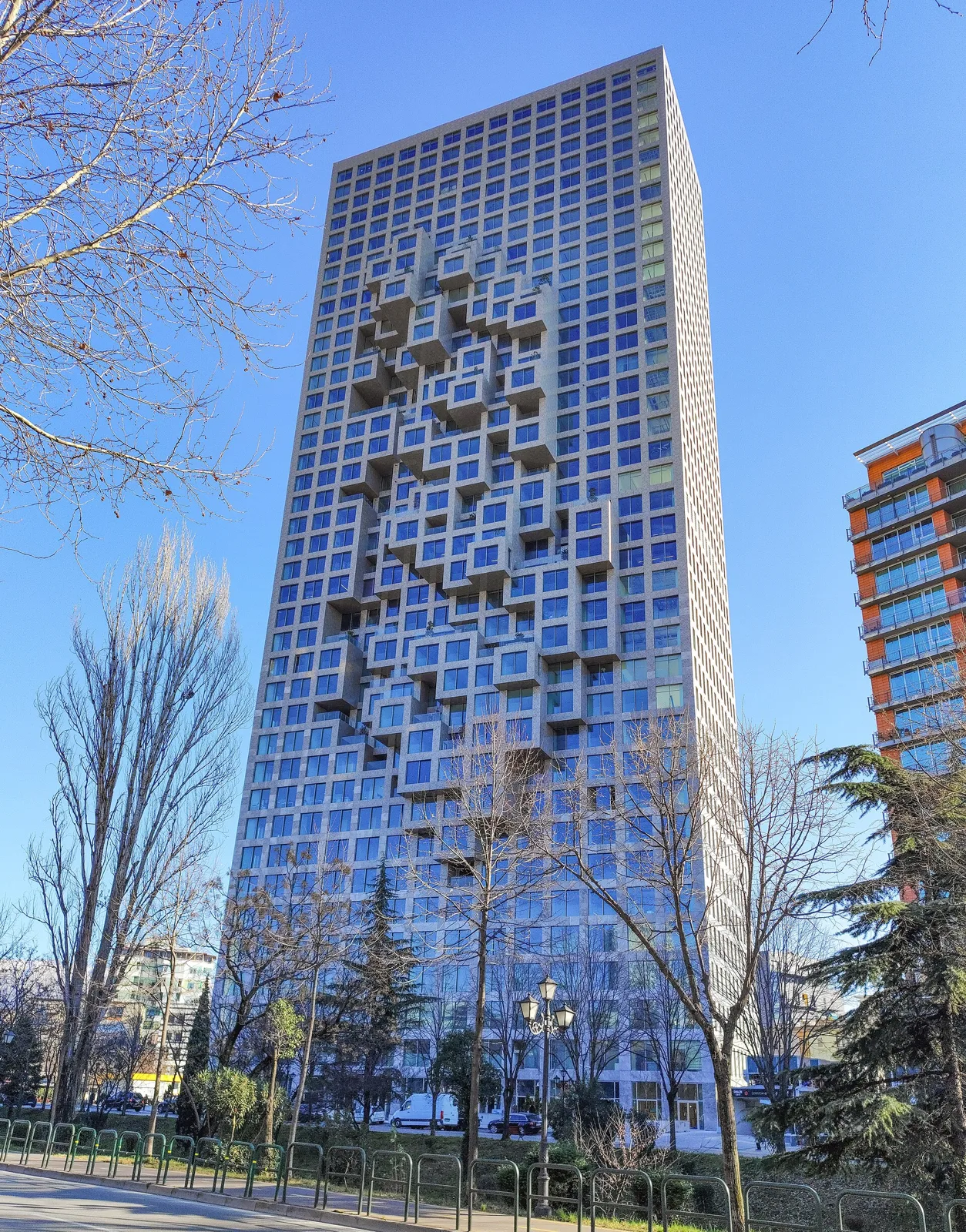 A glass residential tower with a pixelated pattern of recessed balconies on its facade, seen from a tree-lined street