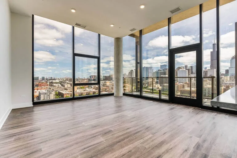 Empty apartment living room with floor-to-ceiling windows, a concrete column, wood-look flooring and a balcony door overlooking a city skyline