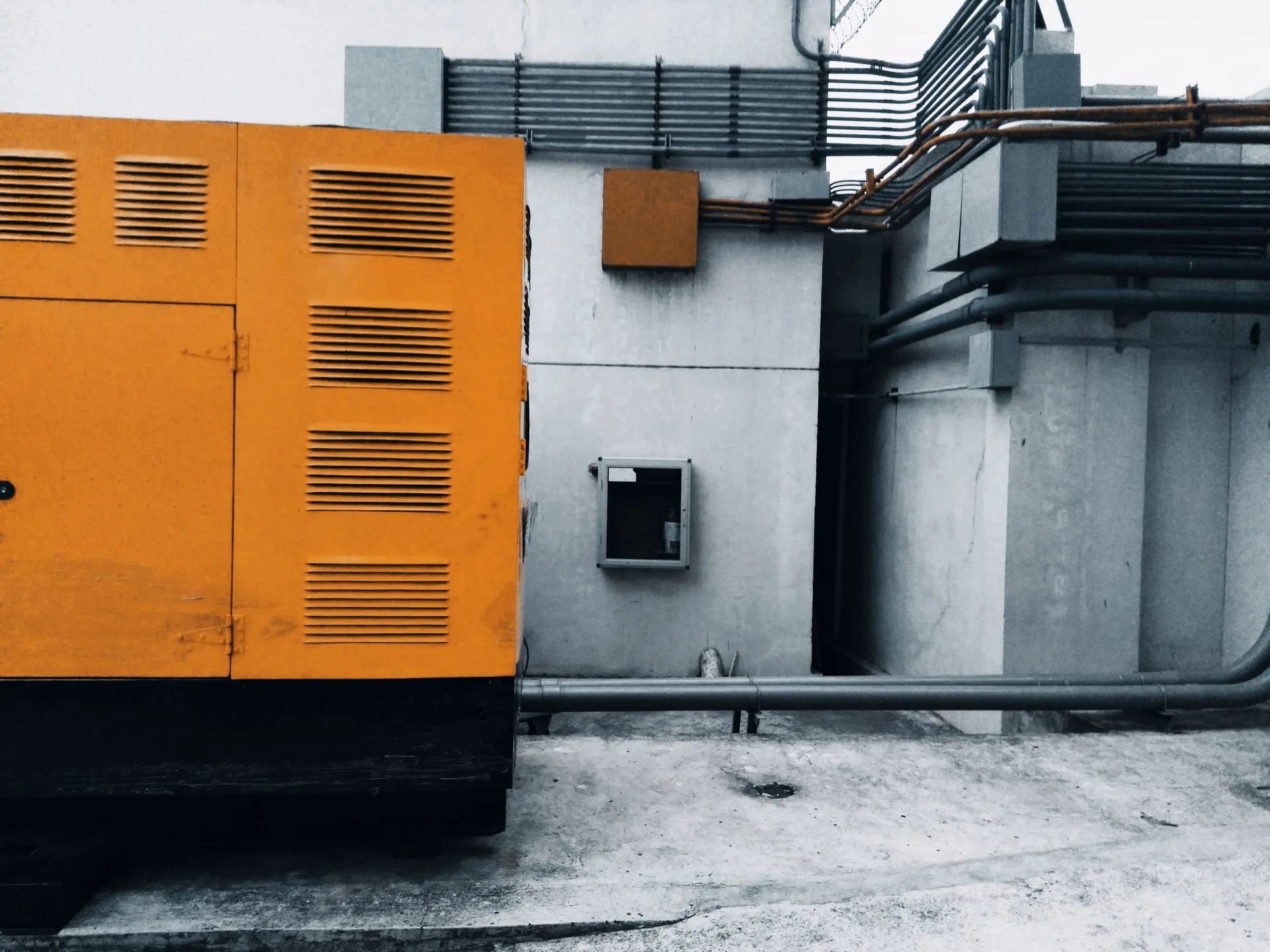 Enclosed standby generator with louvered orange housing beside a building wall and pipework
